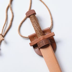 May include: A wooden toy sword featuring a light-colored blade and a handle wrapped in thick tan rope. The sword is held by a brown leather scabbard with brass-colored rivets and a decorative knotwork pattern, attached to a long tan rope neck strap.