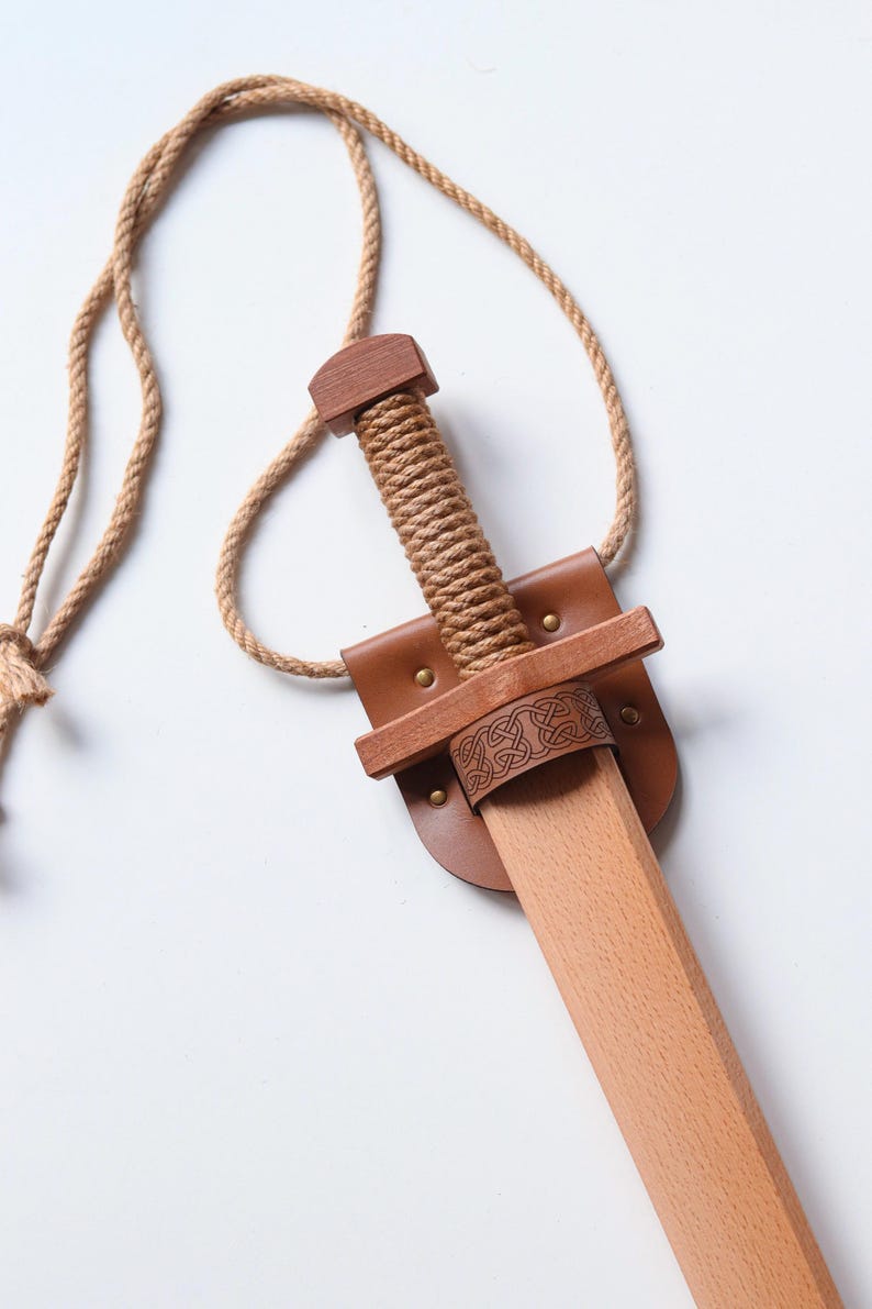 May include: A wooden toy sword featuring a light-colored blade and a handle wrapped in thick tan rope. The sword is held by a brown leather scabbard with brass-colored rivets and a decorative knotwork pattern, attached to a long tan rope neck strap.