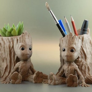 Puede incluir: Dos macetas de madera marrón en forma de Groot. Una maceta tiene una pequeña planta suculenta verde creciendo dentro. La otra maceta tiene un pincel, dos bolígrafos azules, un bolígrafo plateado y un lápiz rojo dentro.
