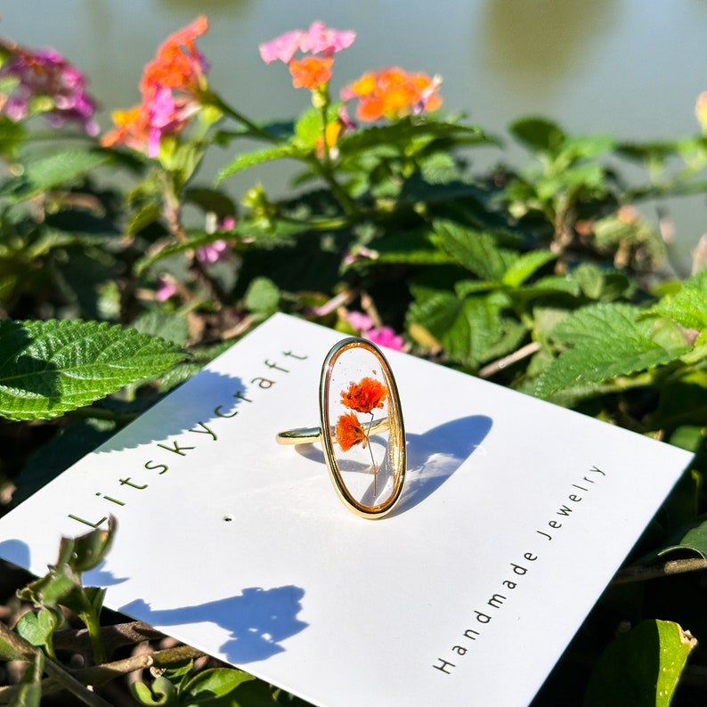 K&ouml;nnte beinhalten: Goldring mit einer ovalen, transparenten Harzfassung, in der gepresste orangefarbene Blumen enthalten sind. Der Ring liegt auf einer wei&szlig;en Karte mit dem Text "Litskycraft Handmade Jewelry".