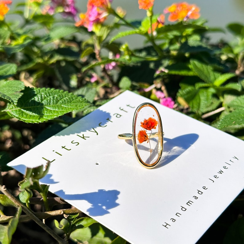 K&ouml;nnte beinhalten: Ein goldener ovaler Ring mit einer klaren Harzfassung, in der eine gepresste orange Blume eingearbeitet ist. Der Ring wird auf einer wei&szlig;en Karte mit dem Text "Litskycraft Handmade Jewelry" pr&auml;sentiert.