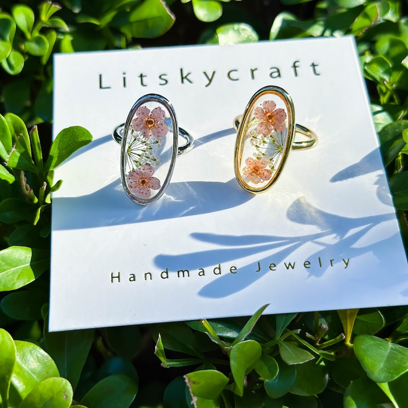 May include: Two oval-shaped rings with delicate pressed flowers encased in clear resin. The rings are on a white card with the text "Litskycraft Handmade Jewelry".