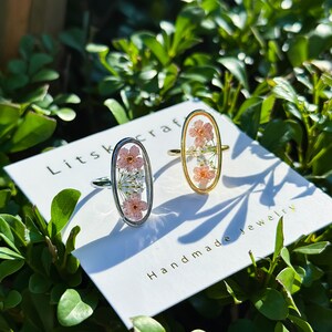 May include: Two oval-shaped rings with delicate pressed flowers encased in clear resin. The rings are silver and gold, and the flowers are pink and white. The rings are displayed on a white card with the text "Litskraf Handmade Jewelry".