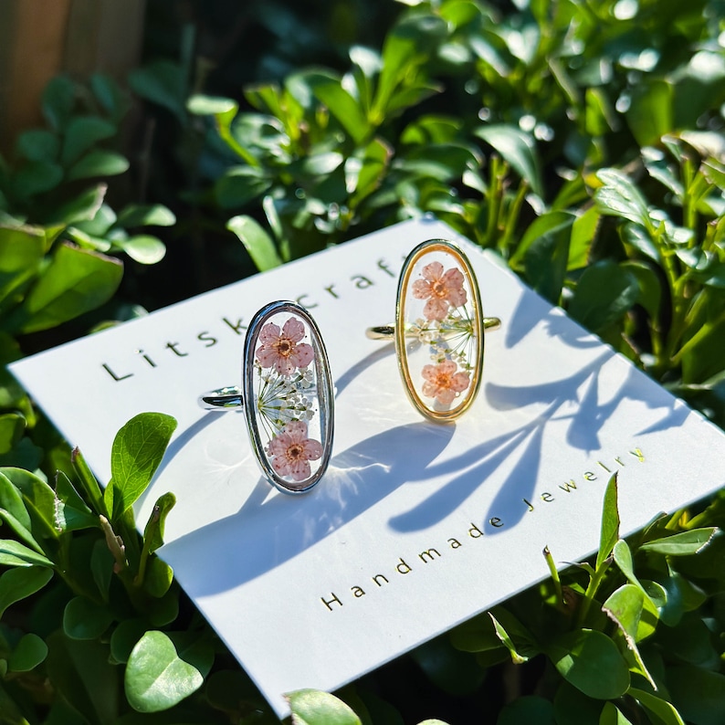 May include: Two oval-shaped rings with delicate pressed flowers encased in clear resin. The rings are silver and gold, and the flowers are pink and white. The rings are displayed on a white card with the text "Litskraf Handmade Jewelry".