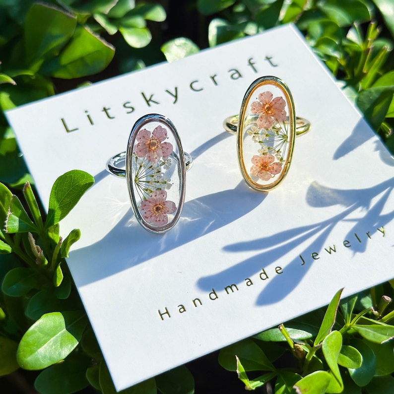 May include: Two oval-shaped rings with delicate pressed flowers encased in clear resin. The rings are on a white card with the text "Litsky craft Handmade Jewelry".