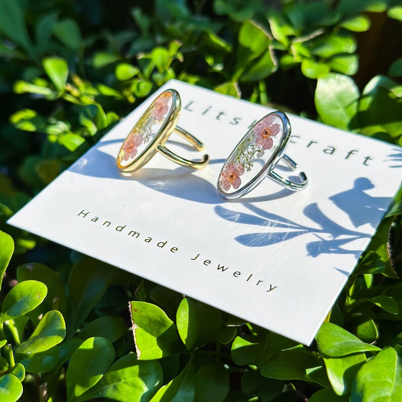 May include: Two oval-shaped rings with delicate pressed flowers encased in clear resin. The ring on the left has a gold band, while the ring on the right has a silver band. The rings are displayed on a white card with the text "Litscraft Handmade Jewelry".