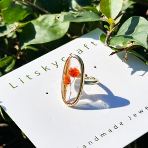 K&ouml;nnte beinhalten: Goldring mit einer ovalen Harzfassung, in der eine gepresste orange Blume eingearbeitet ist. Der Ring liegt auf einer wei&szlig;en Karte mit dem Text "Litskycraft" und "Handgemachter Schmuck".