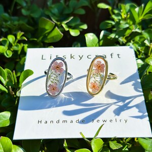 May include: Two oval-shaped rings with delicate pink flowers pressed inside. The rings are on a white card with the text "Litskycraft Handmade Jewelry".