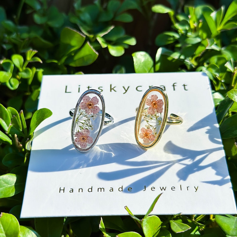 May include: Two oval-shaped rings with delicate pink flowers pressed inside. The rings are on a white card with the text "Litskycraft Handmade Jewelry".