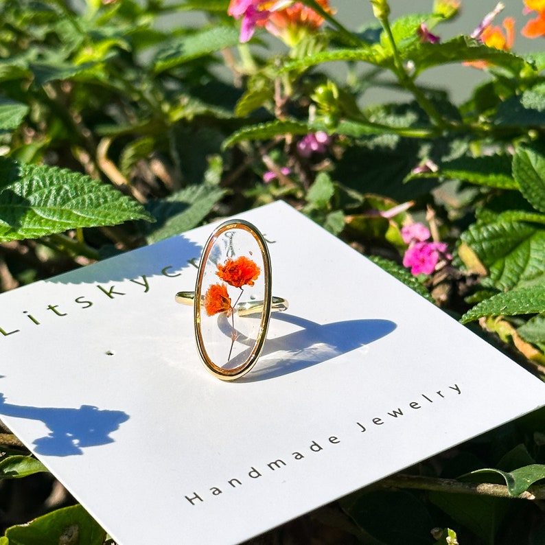 K&ouml;nnte beinhalten: Ein goldener Ring mit einer klaren ovalen Fassung, in der eine gepresste orange Blume eingefasst ist. Der Ring wird auf einer wei&szlig;en Karte mit dem Text "Litsky Craft Handmade Jewelry" pr&auml;sentiert.