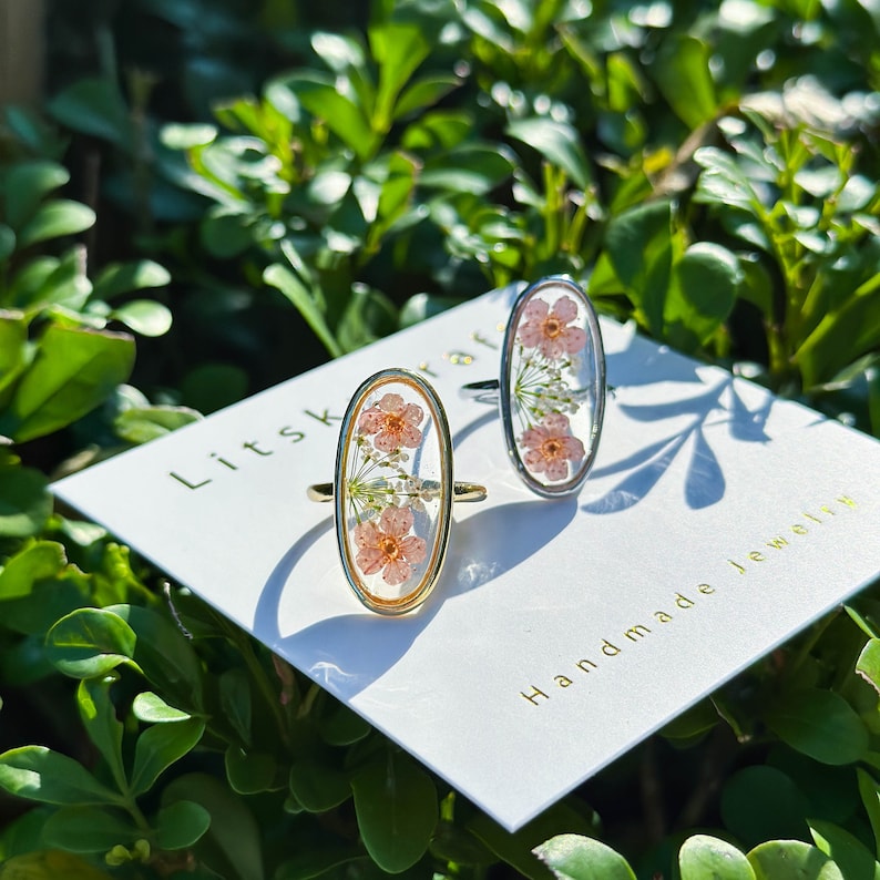 May include: Two oval-shaped rings with delicate pink flowers pressed inside. The rings are on a white card with the text "Litskraft Handmade Jewelry".