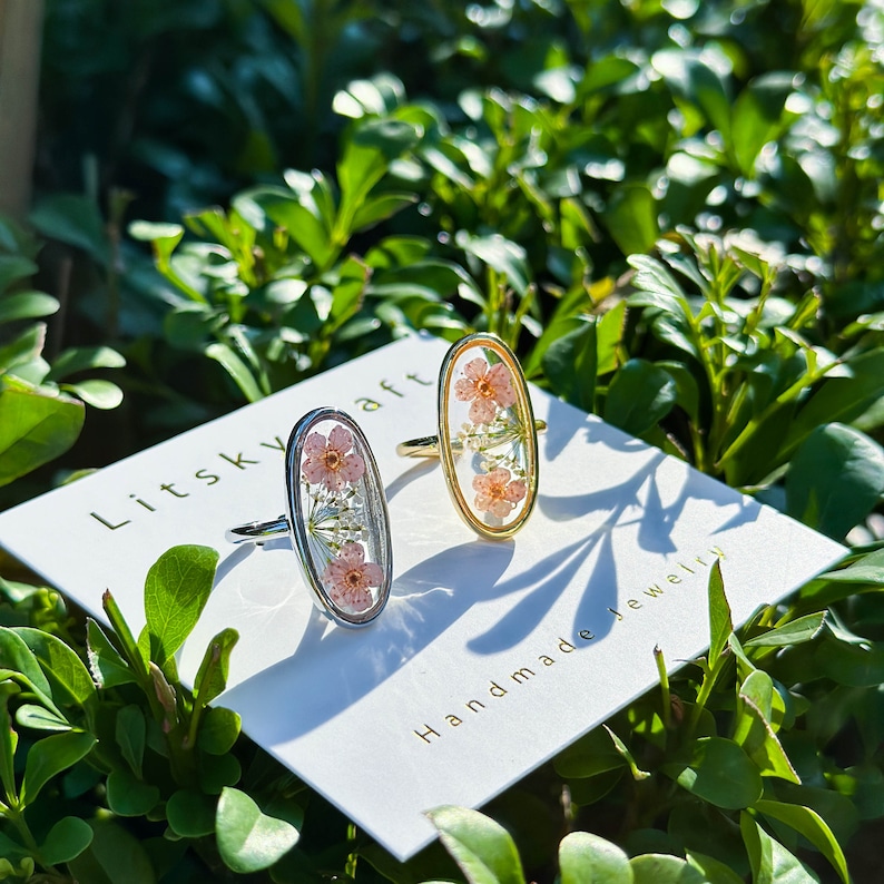 May include: Two oval-shaped rings with delicate pressed flowers inside. The rings are silver and gold and are displayed on a white card with the text "Litskraft Handmade Jewelry".