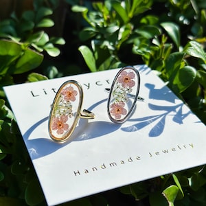 May include: Two oval-shaped rings with delicate pink and white pressed flowers encased in clear resin. The rings are set in gold and silver metal.