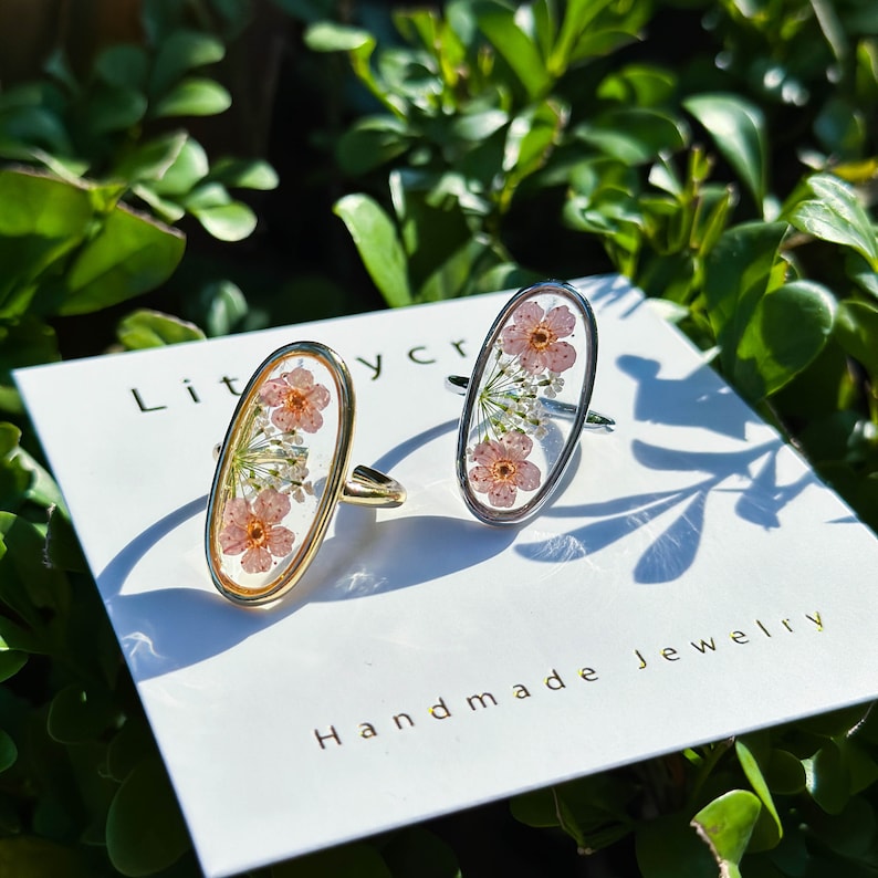 May include: Two oval-shaped rings with delicate pink and white pressed flowers encased in clear resin. The rings are set in gold and silver metal.