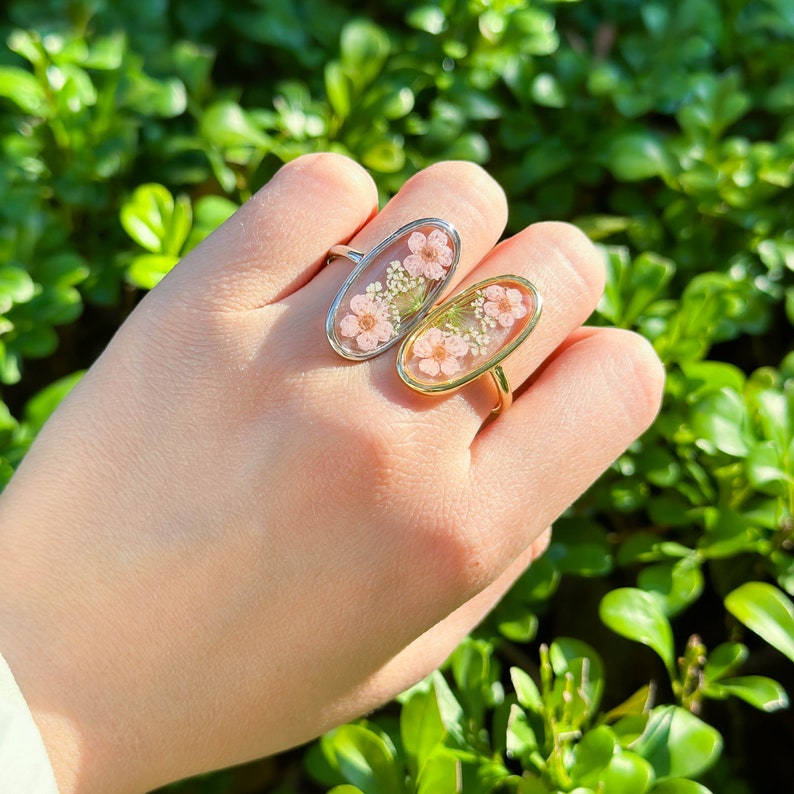 May include: Two oval-shaped rings with delicate floral designs encased in clear resin. One ring has a silver band and the other has a gold band.