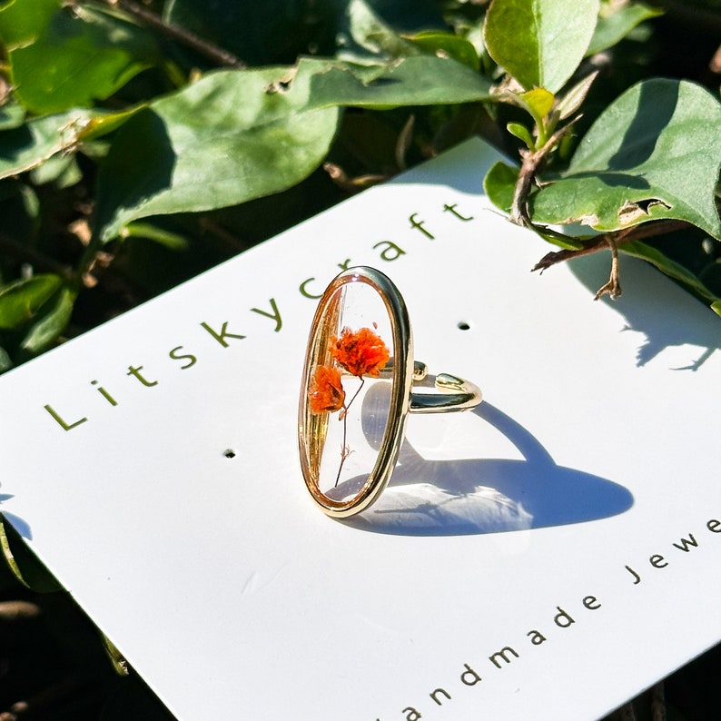 K&ouml;nnte beinhalten: Ein goldener Ring mit einer ovalen Harzfassung, in der eine gepresste orange Blume eingearbeitet ist. Der Ring wird auf einer wei&szlig;en Karte mit dem Text "Litskycraft" und "Handgefertigte Schmuckst&uuml;cke" pr&auml;sentiert.