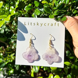 Könnte beinhalten: Ein Paar zarte Ohrringe mit gepressten lila Blüten, die in klarem Harz gefasst sind. Die Ohrringe hängen an goldfarbenen Haken und haben ein schlichtes, elegantes Design. Die Karte, auf der sie ausgestellt sind, trägt den Schriftzug "Litskycraft".