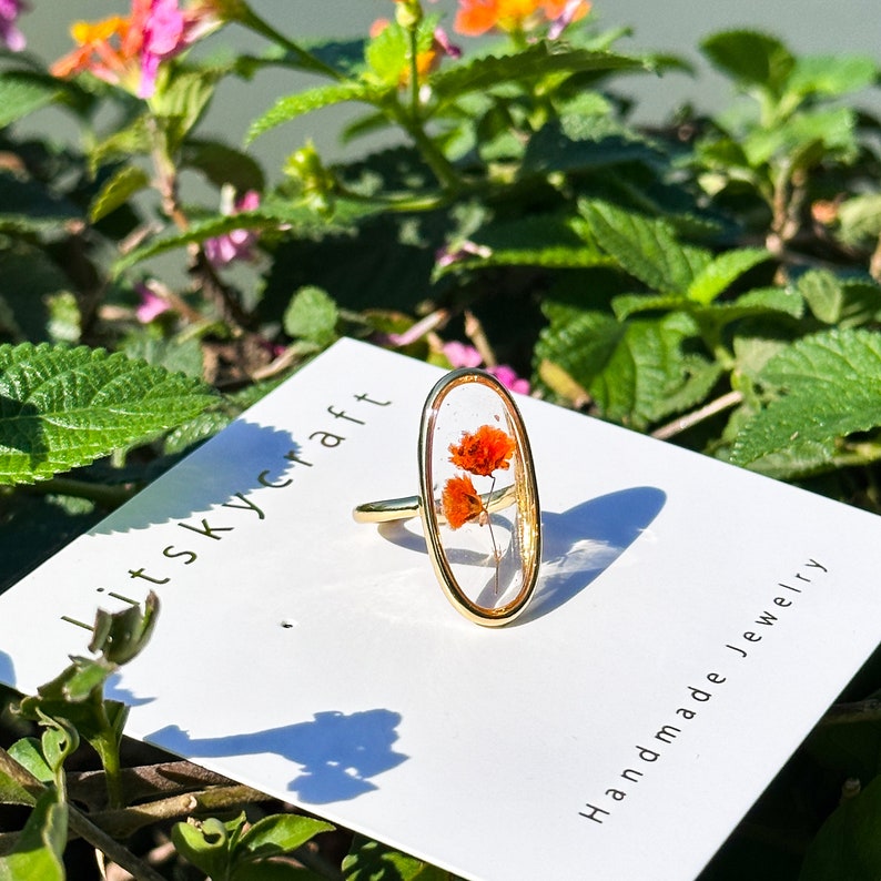 K&ouml;nnte beinhalten: Ein goldener Ring mit einer ovalen, transparenten Harzfassung, in der sich zwei kleine, getrocknete orangefarbene Bl&uuml;ten befinden. Der Ring wird auf einer wei&szlig;en Karte mit dem Text "lit sky craft" und "Handmade Jewelry" pr&auml;sentiert.