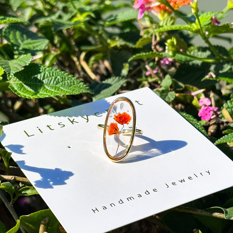 K&ouml;nnte beinhalten: Ein goldener ovaler Ring mit einer klaren Harzfassung, in der gepresste orangefarbene Blumen eingearbeitet sind. Der Ring wird auf einer wei&szlig;en Karte mit dem Text "Litsky Craft Handmade Jewelry" pr&auml;sentiert.