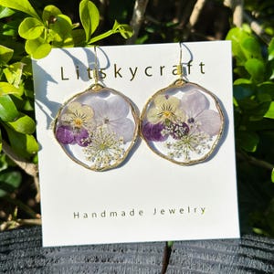 Puede incluir: Un par de pendientes dorados hechos a mano con un diseño circular. Cada pendiente presenta un círculo relleno de resina transparente con flores prensadas moradas, blancas y amarillas. Texto: "Litskycraft Handmade Jewelry".