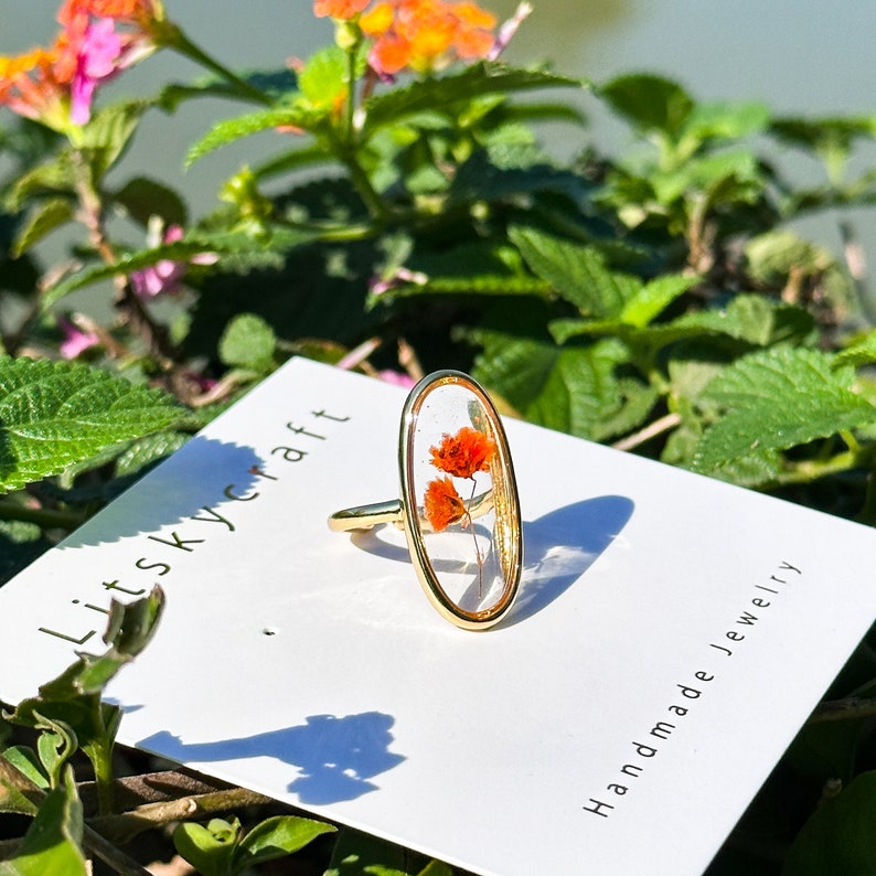 K&ouml;nnte beinhalten: Ein goldener Ring mit einer klaren ovalen Fassung, in der sich zwei kleine, gepresste orangefarbene Bl&uuml;ten befinden. Der Ring wird auf einer wei&szlig;en Karte mit dem Text "Litskycraft Handmade Jewelry" pr&auml;sentiert.