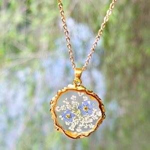 Puede incluir: Un collar de cadena dorada con un colgante redondo que presenta una delicada disposición de flores azules y blancas prensadas encapsuladas en resina transparente.