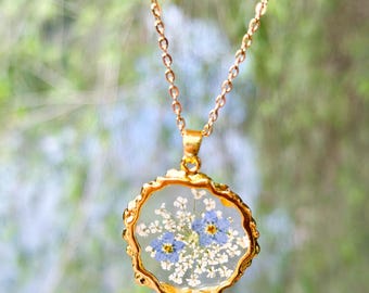 May include: A gold chain necklace with a round pendant featuring a delicate arrangement of pressed blue and white flowers encased in clear resin.