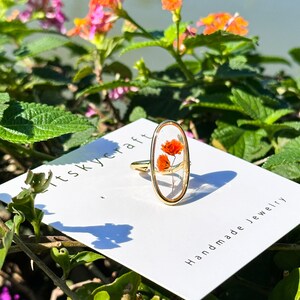 K&ouml;nnte beinhalten: Ein goldener Ring mit einer klaren ovalen Harzfassung, in der zwei kleine, getrocknete orangefarbene Blumen zu sehen sind. Der Ring wird auf einer wei&szlig;en Karte mit dem Text "it's skycraft handmade jewelry" pr&auml;sentiert.