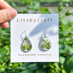 Puede incluir: Un par de pendientes en forma de lágrima con ganchos plateados. Cada pendiente presenta una lágrima de resina transparente con una flor verde prensada en su interior. Los pendientes se muestran en una tarjeta blanca con el texto "Litskycraft Handmade Jewelry".