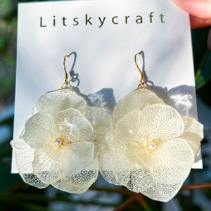 May include: A pair of delicate white flower earrings with gold hooks and a pearl accent in the centre of each flower.