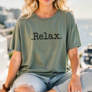 May include: A woman wearing a green t-shirt with the text ".Relax." printed on it. She is wearing ripped jeans and sitting on a wooden surface.