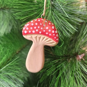 Può includere: Un ornamento natalizio a forma di fungo rosso e bianco. L'ornamento ha un cappello rosso con pois bianchi e un gambo beige. È appeso a un filo di spago naturale su uno sfondo di aghi di pino verdi.