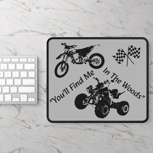 May include: A gray mousepad with black outlines featuring two black and white illustrations of dirt bikes and the text "You'll Find Me In The Woods".