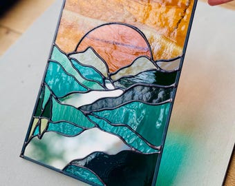 Può includere: Una finestra in vetro colorato che rappresenta un tramonto su una catena montuosa con una gamma di colori verde acqua e verde.