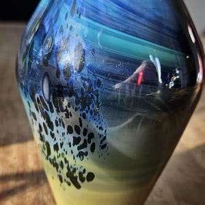 Peut inclure: Un vase en verre artisanal au design unique. Le vase présente un mélange de teintes bleues, vertes et jaunes, avec des accents tachetés foncés. Il a un dessus évasé et un corps arrondi, idéal pour exposer des fleurs ou comme pièce décorative.