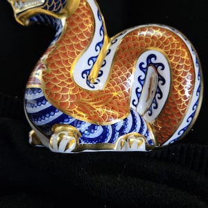 Può includere: Una statuetta in porcellana di un drago con squame blu, dorate e arancioni. Il drago è rannicchiato in una posizione di riposo con la testa girata di lato.
