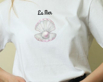 Può includere: Maglietta bianca con una conchiglia rosa e bianca con una perla al suo interno. Il testo "La Mer" è stampato sopra la conchiglia.