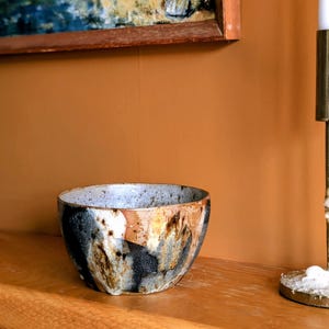 Op de afbeelding: Een handgemaakte keramische kom met een uniek, getextureerd oppervlak. De kom heeft een mix van kleuren, waaronder zwart, bruin en wit, waardoor een gemarmerd effect ontstaat. Hij staat op een houten oppervlak en toont zijn artistieke ontwerp. Een decoratief item voor in huis.