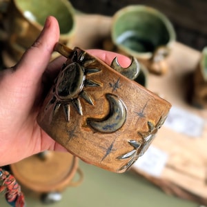 May include: A cylindrical ceramic mug with a tan, speckled glaze featuring raised, dark-toned relief designs of a sun, a crescent moon, and small stars. The surface has a glossy finish, and the celestial motifs are distributed around the exterior.