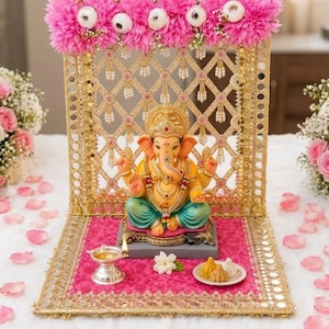 May include: A Ganesha statue sits on a pink fabric base in front of a gold lattice screen decorated with pink flowers. The statue features yellow skin, teal trousers, and gold jewellery. A silver oil lamp and a small plate of modak sweets are placed nearby.