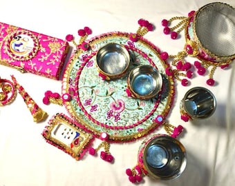 May include: A traditional Indian wedding ceremony set with a round, turquoise and pink embroidered tray, four silver bowls, a pink and gold box, a pink and gold bangle, a pink and gold cone, and a pink and gold phone case.