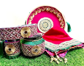 May include: A set of traditional Indian wedding ceremony items, including a round silver tray with a pink velvet base and gold accents, three silver bowls with pink and gold trim, and a red silk scarf with gold and green embroidery.