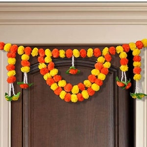 Può includere: Una ghirlanda di fiori gialli e arancioni, nota anche come appendino per porta, è realizzata con pompon di stoffa e infilata con filo bianco. La ghirlanda è decorata con foglie verdi ed è appesa a una porta in legno marrone.