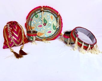 May include: A collection of decorative items, including an embroidered plate with a light blue background and paisley designs, a red and gold drawstring bag, and two small metal cups. The items are decorated with tassels and beaded garlands.