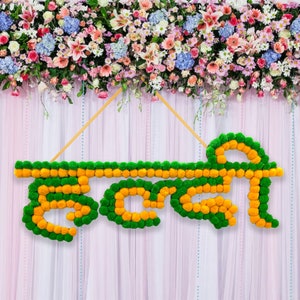 May include: A floral arrangement spelling out the word "Happy" in Hindi using yellow and green marigold flowers. The flowers are strung together to form the letters. The arrangement is hanging from a white sheer curtain backdrop.