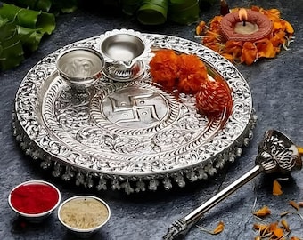 Peut inclure: Un thali en argent, ou plateau cérémoniel, avec des motifs floraux complexes et un symbole swastika au centre. Le thali est entouré de petits bols, d'un diya et de fleurs de souci.