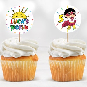 May include: Two cupcakes with colorful toppers. One topper features a cartoon sun with the text "Luca's World". The other topper features a cartoon superhero with the number "5".