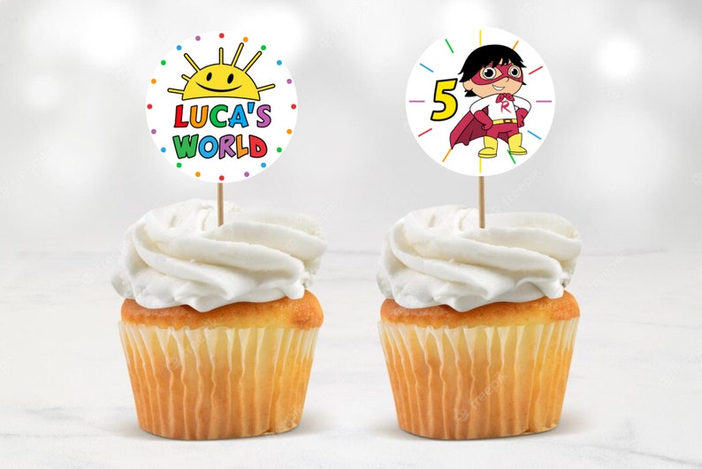 May include: Two cupcakes with colourful toppers. One topper features a cartoon sun with the text "Luca's World". The other topper features a cartoon superhero with the number "5".