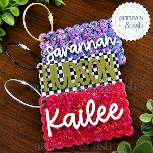 May include: Three rectangular luggage tags with scalloped edges and metal cable loops. The top tag is purple glitter with the white name Savannah. The middle tag has a black and white checkerboard pattern with the olive green name Hudson. The bottom tag is red glitter with the white name Kailee.