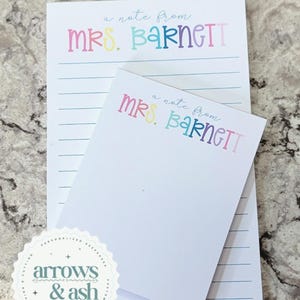 May include: Two white notepads, one large and one small, with the text "a note from MRS. BARNETT" in colorful letters. The larger notepad has blue horizontal lines. The smaller notepad is blank. A circular logo with the text "arrows & ash" is in the lower left corner.